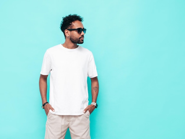 Handsome smiling hipster model Sexy unshaven man dressed in white summer tshirt and jeans clothes Fashion male posing near blue wall in studio Isolated In sunglasses