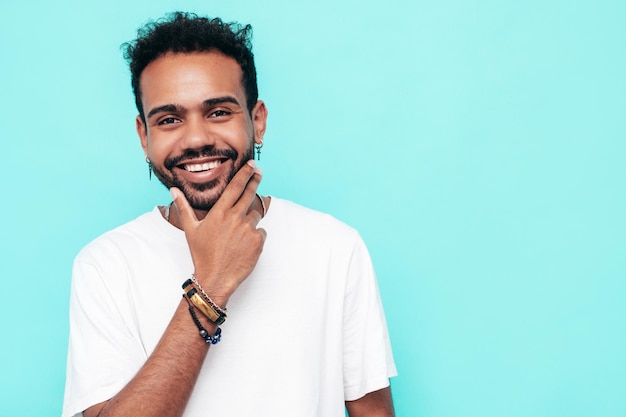 Handsome smiling hipster model sexy unshaven man dressed in white summer tshirt and jeans clothes fashion male posing near blue wall in studio isolated