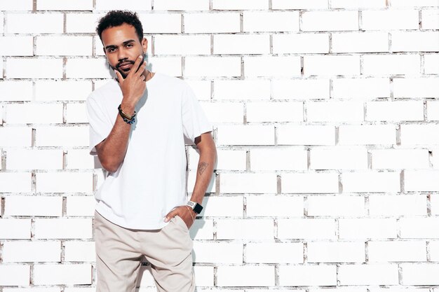 Handsome smiling hipster model Sexy unshaven man dressed in white summer tshirt and jeans clothes Fashion male posing near white brick wall in studio at sunny day Isolated