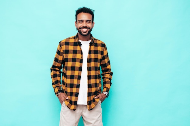 Handsome smiling hipster model Sexy unshaven man dressed in yellow summer shirt and jeans clothes Fashion male posing near blue wall in studio Cheerful and happy Isolated