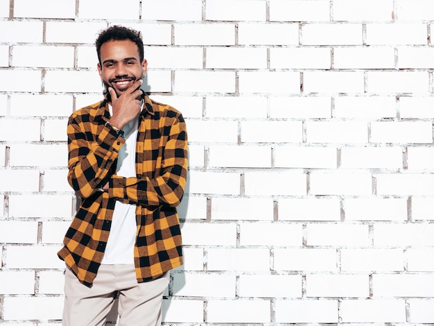 Handsome smiling hipster model Sexy unshaven man dressed in yellow summer shirt and jeans clothes Fashion male posing near white brick wall in studio at sunny day