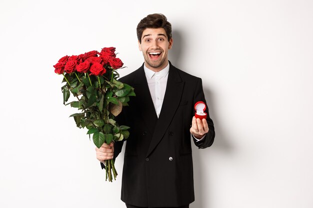 Handsome smiling man in black suit, holding roses and engagement ring, making a proposal to marry him, standing against white background.