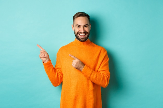 Handsome smiling man pointing fingers left, showing your logo, standing in winter orange sweater, turquoise wall.