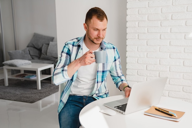 Handsome smiling man in shirt sitting in kitchen at home at table working online on laptop from home