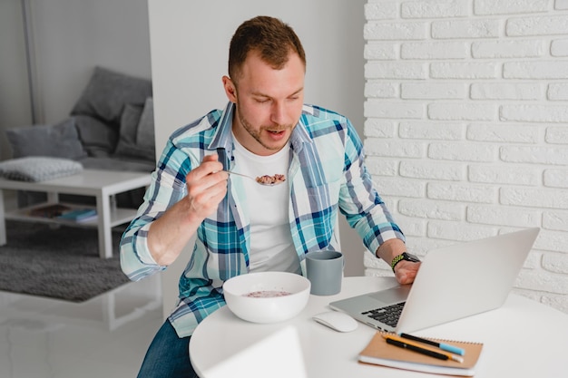 Bell'uomo sorridente in camicia seduto in cucina a casa al tavolo che lavora online sul laptop