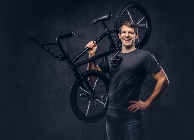 Handsome smiling man in t-shirt and jeans holding BMX bicycle on his shoulder on a dark background.