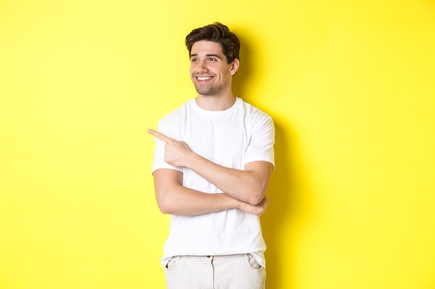 Handsome smiling man in white clothes, looking and pointing finger left at banner, standing over yellow background.