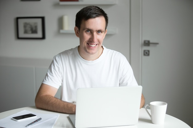 Handsome smiling man at the white desk working with laptop