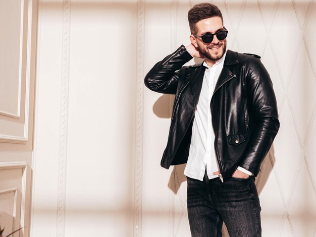 Handsome smiling model Sexy stylish man in biker leather jacket and black jeans Fashion hipster male posing near white wall in studio interior at sunny day Shadow from window in sunglasses