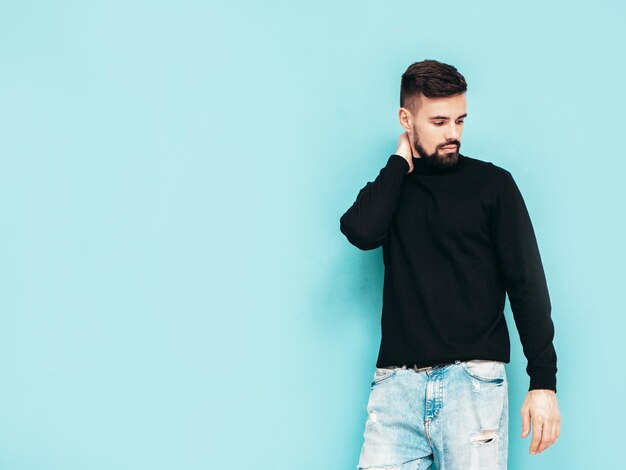 잘 생긴 웃는 모델 섹시 한 세련 된 남자 검은 터틀넥 스웨터와 청바지를 입고 패션 힙 스터 남성 스튜디오에서 파란색 벽 근처 포즈 절연