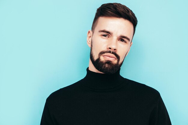 Handsome smiling model Sexy stylish man dressed in black turtleneck sweater and jeans Fashion hipster male posing near blue wall in studio Isolated