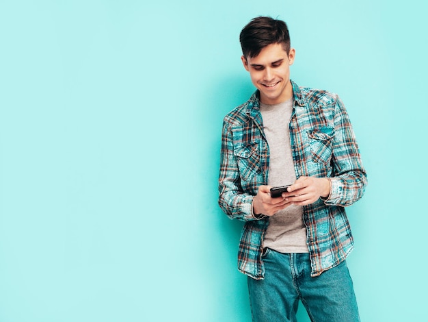 Modello sorridente bello uomo alla moda sexy vestito con camicia a scacchi e jeans moda uomo hipster in posa vicino al blu in studio isolato holding smartphone guardando lo schermo del cellulare utilizzando app