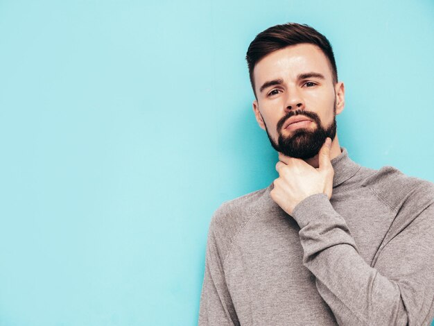 Handsome smiling model Sexy stylish man dressed in grey turtleneck sweater and jeans Fashion hipster male posing near blue wall in studio Isolated