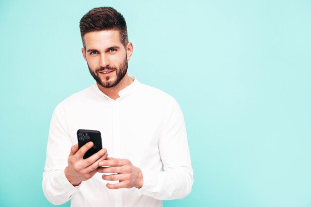 Handsome smiling modelSexy stylish man dressed in shirt and jeans Fashion hipster male posing near blue wall in studio Holding smartphone Looking at cellphone screen Using apps