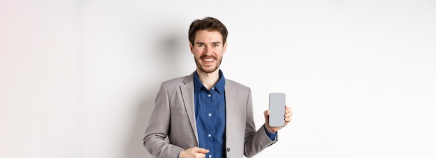 Free photo handsome smiling salesman in suit showing empty smartphone screen demonstrate an app standing on whi