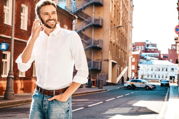 Handsome smiling stylish hipster lambersexual modelModern man dressed in white shirt Fashion male posing on the street background in sunglasses Outdoors at sunset Using smartphoneTalking