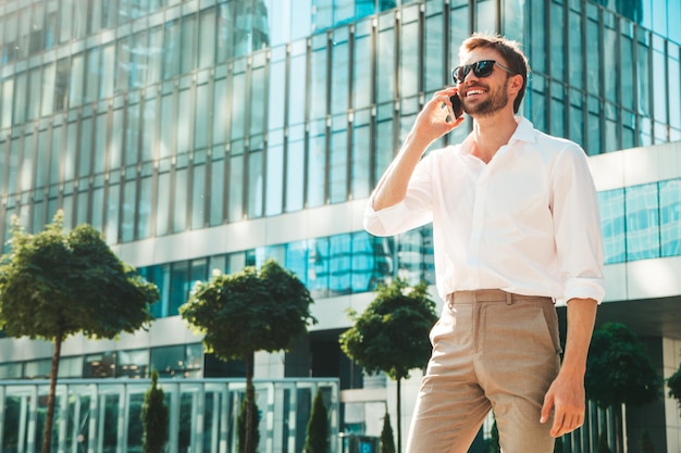 Handsome smiling stylish hipster lambersexual modelModern man dressed in white shirt Fashion male posing on the street background in sunglasses Outdoors at sunset Using smartphoneTalking