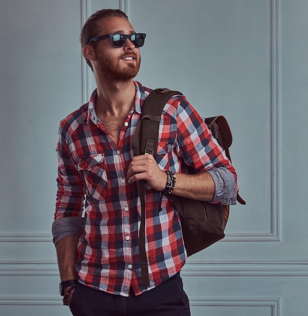 Free photo a handsome smiling stylish redhead traveler in a flannel shirt and sunglasses with a backpack, posing in a studio against a white wall.