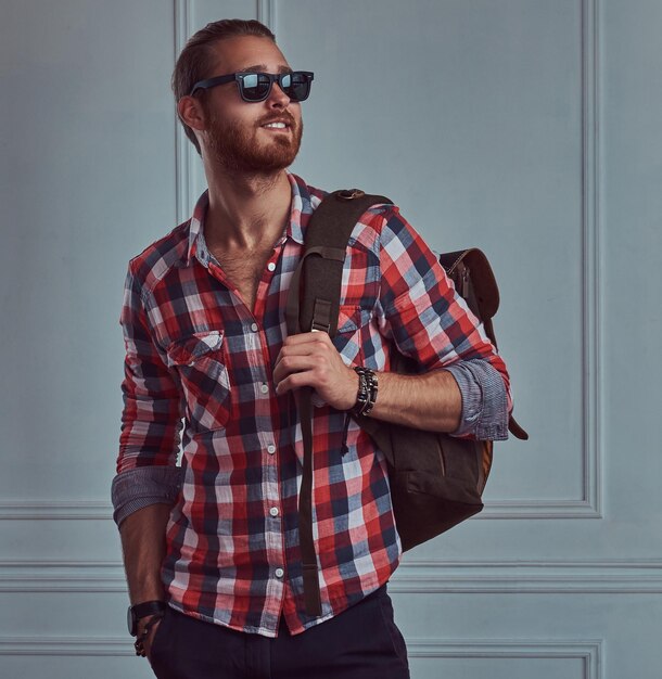 A handsome smiling stylish redhead traveler in a flannel shirt and sunglasses with a backpack, posing in a studio against a white wall.
