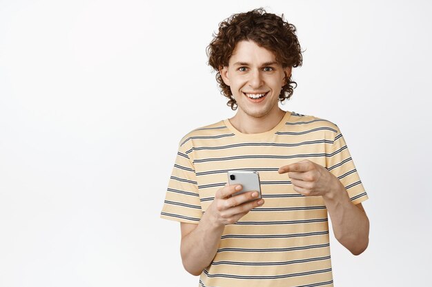 Красивый улыбающийся подросток, указывающий на свой мобильный телефон, говорит о приложении или интернет-магазине, стоящем на белом фоне