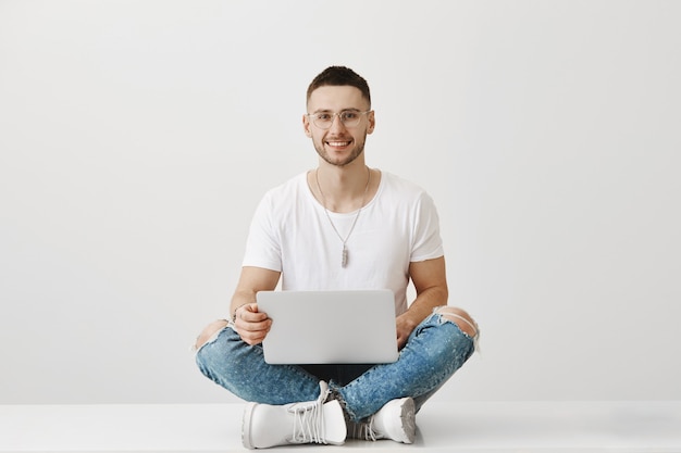 Foto gratuita bel ragazzo giovane sorridente con gli occhiali in posa con il suo computer portatile