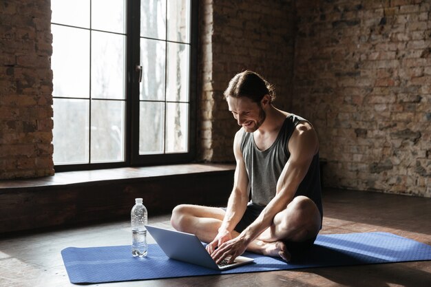 Handsome strong sportsman sitting near bottle of water using laptop