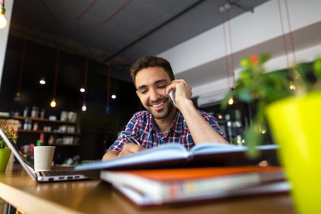 Handsome student speaking over mobile or smart phone while studying over classes in restaurant or cafe.