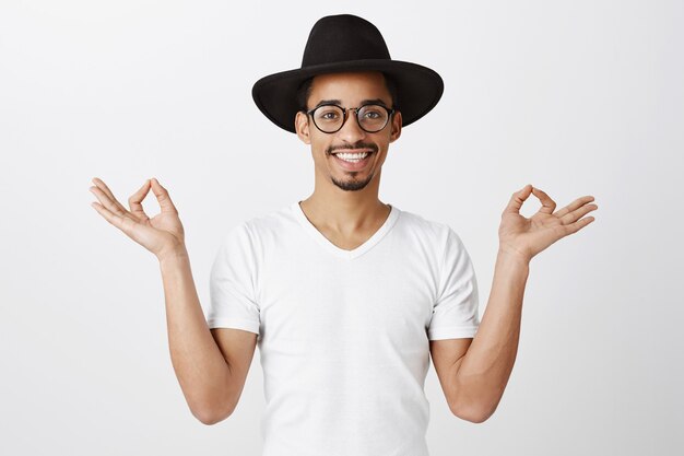 Handsome stylish african american guy in hipster hat and glasses meditating, practice yoga