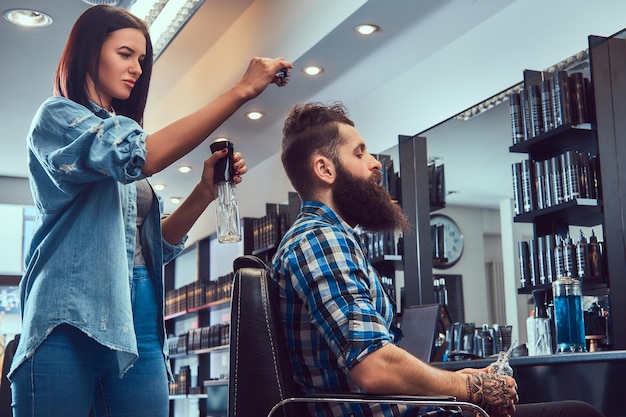 Handsome stylish bearded male with a tattoo on arm dressed in a flannel shirt holding juice while getting a haircut.