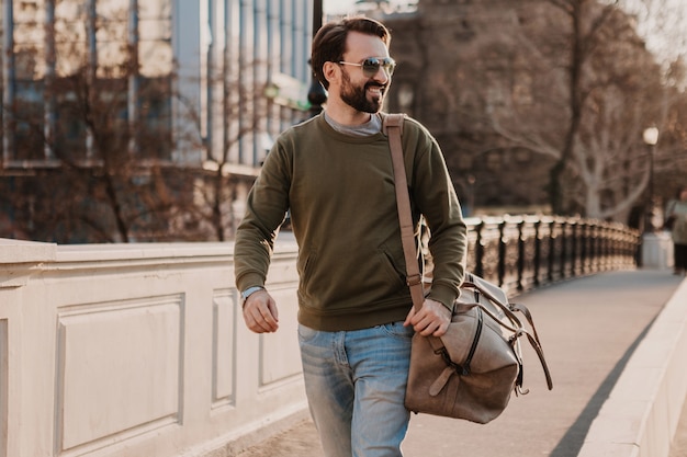 Free photo handsome stylish bearded man walking in city street with leather travel bag wearing sweatshirt and sunglasses, urban style trend, sunny day, confident and smiling