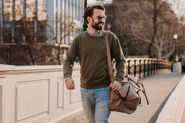 Handsome stylish bearded man walking in city street with leather travel bag wearing sweatshirt and sunglasses, urban style trend, sunny day, confident and smiling