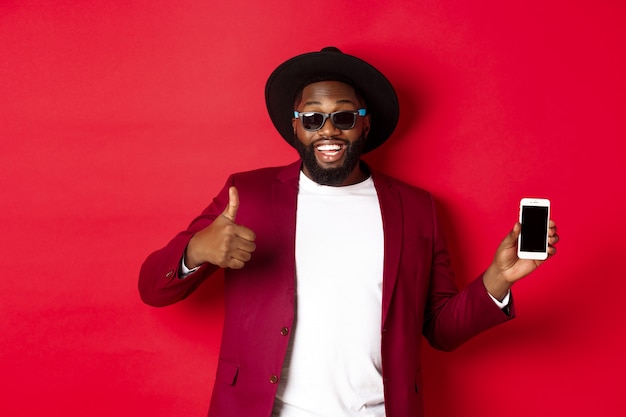 Handsome and stylish Black man showing phone screen and thumb up at camera, recommending online store app, red background