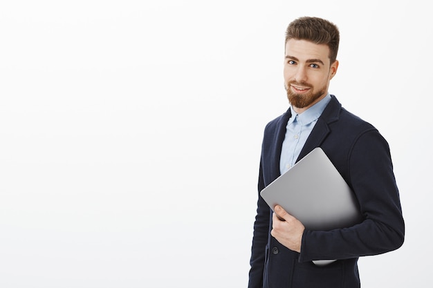 Handsome and stylish businessman knows how business works. Successful and determined good-looking man in suit holding laptop computer in hand and gazing self-assured against grey wall