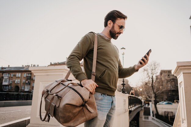 Handsome stylish hipster man walking in city street with leather bag using phone navigation application, travel wearing sweatshirt and sunglasses, urban style trend