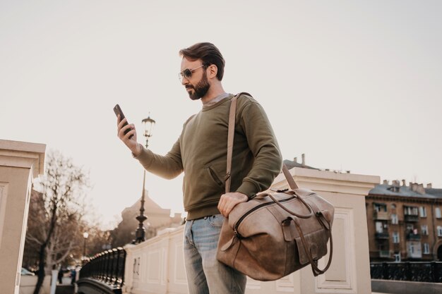 Handsome stylish hipster man walking in city street with leather bag using phone, travel wearing sweatshirt and sunglasses, urban style trend, sunny day