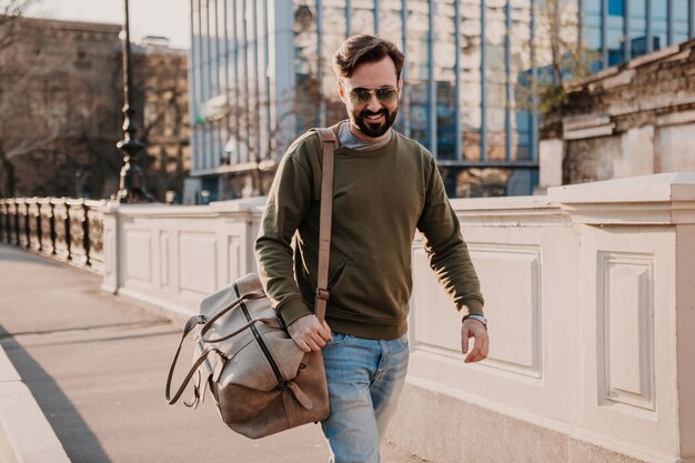 Handsome stylish hipster man walking in city street with leather bag wearing sweatshot and sunglasses, urban style trend, sunny day, smiling happy traveler