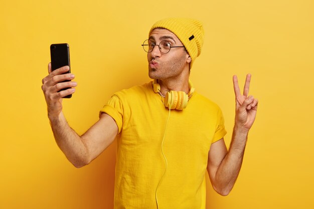 Handsome stylish hipster takes selfie, makes peace gesture, keeps lips folded, holds smart phone