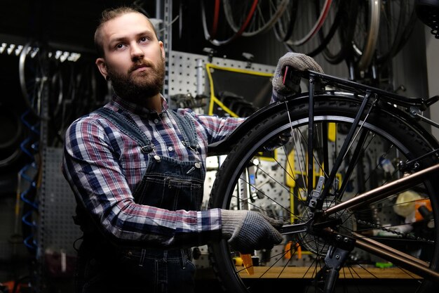 フランネルシャツとジーンズのつなぎ服を着て、修理店で自転車のホイールを操作しているハンサムなスタイリッシュな男性。レンチを使用している作業員が、ワークショップで自転車にホイールを取り付けます。