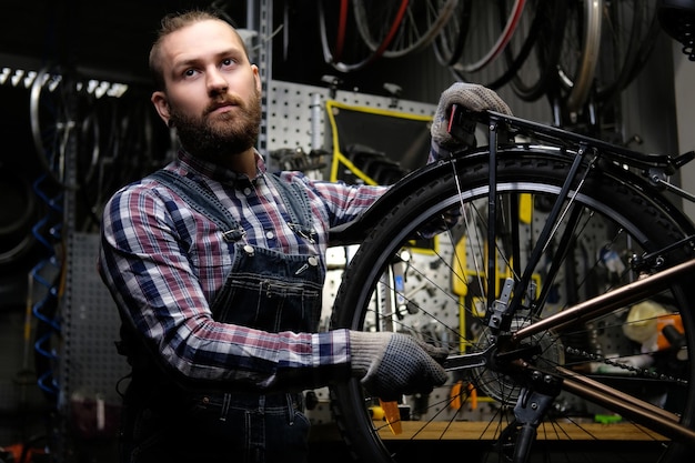 Красивый стильный мужчина в фланелевой рубашке и джинсовом комбинезоне, работающий с велосипедным колесом в ремонтной мастерской. Рабочий с помощью гаечного ключа устанавливает колесо на велосипед в мастерской.