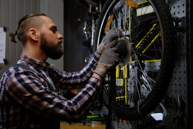 Free photo a handsome stylish male wearing a flannel shirt and jeans coverall, working with a bicycle wheel in a repair shop.