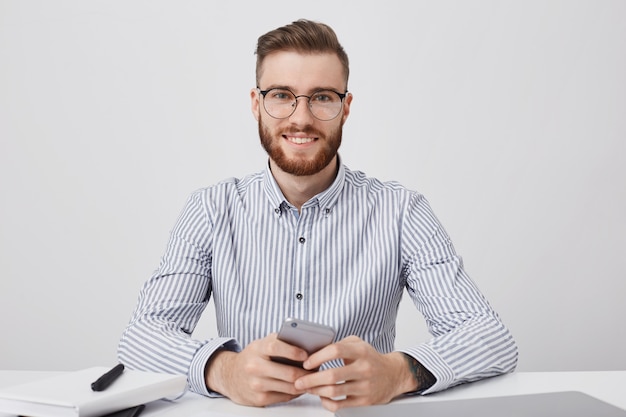 Handsome stylish man dressed formally, sits at working desk, uses smart phone for reading news online