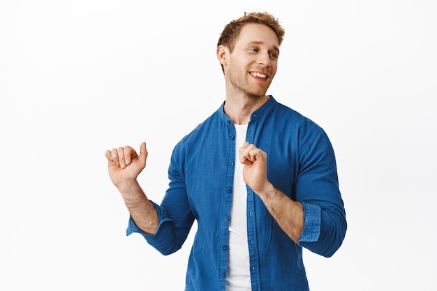 Handsome stylish redhead guy dancing, having fun and smiling, relaxing on dance floor, looking aside with satisfied chill face, standing over white wall