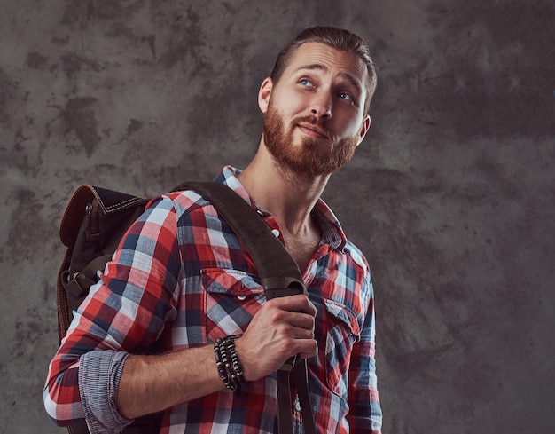 Foto gratuita un bel viaggiatore rosso alla moda in una camicia di flanella con uno zaino, in posa in uno studio su sfondo grigio.