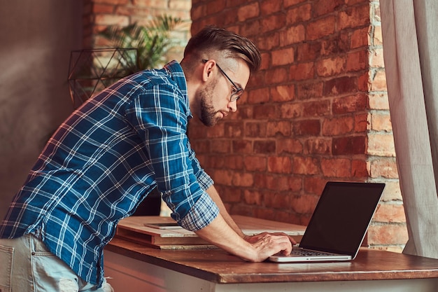 Studente bello ed elegante con una camicia di flanella che lavora su un computer portatile in una stanza con interni soppalcati.