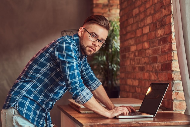 Studente bello ed elegante con una camicia di flanella che lavora su un computer portatile in una stanza con interni soppalcati.