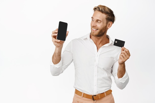 Handsome stylish young man looking at mobile phone screen app interface showing credit card white background