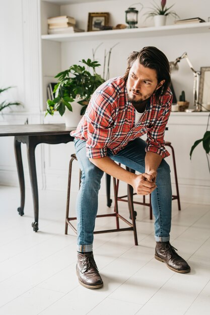 Handsome stylish young man sitting on stool at home looking away