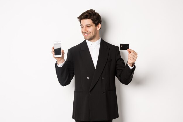 Handsome successful businessman, looking at smartphone screen and showing credit card, standing in black suit against white background