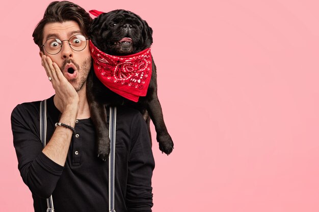 Handsome surprised young unshaven male stratup designer holds black dog on shoulder, keeps hand on cheek, forgets about something, stands against pink wall with copy space. Friendship concept