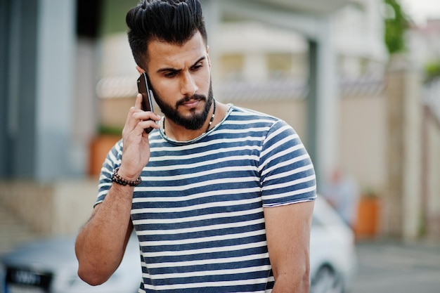 벗은 셔츠를 입은 잘생긴 키 큰 아라비아 수염 남자 모델은 야외에서 차에 대고 포즈를 취했다. 세련된 아랍 남자 이슬람 사업가가 휴대전화에서 말하는